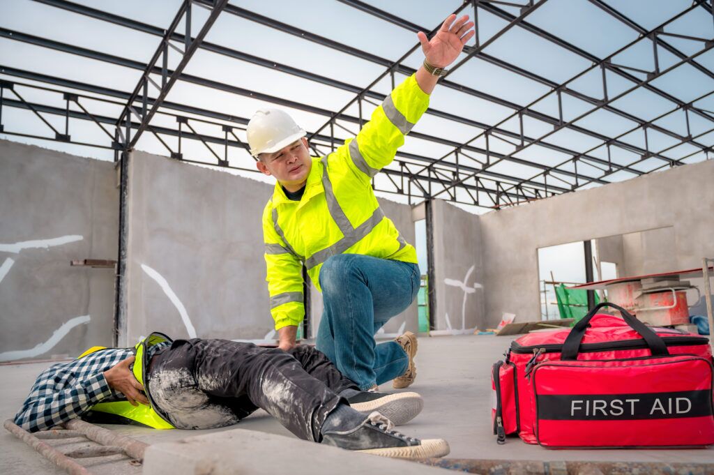Worker kneeling to treat an injured coworker’s leg with a first aid kit nearby on a construction site.