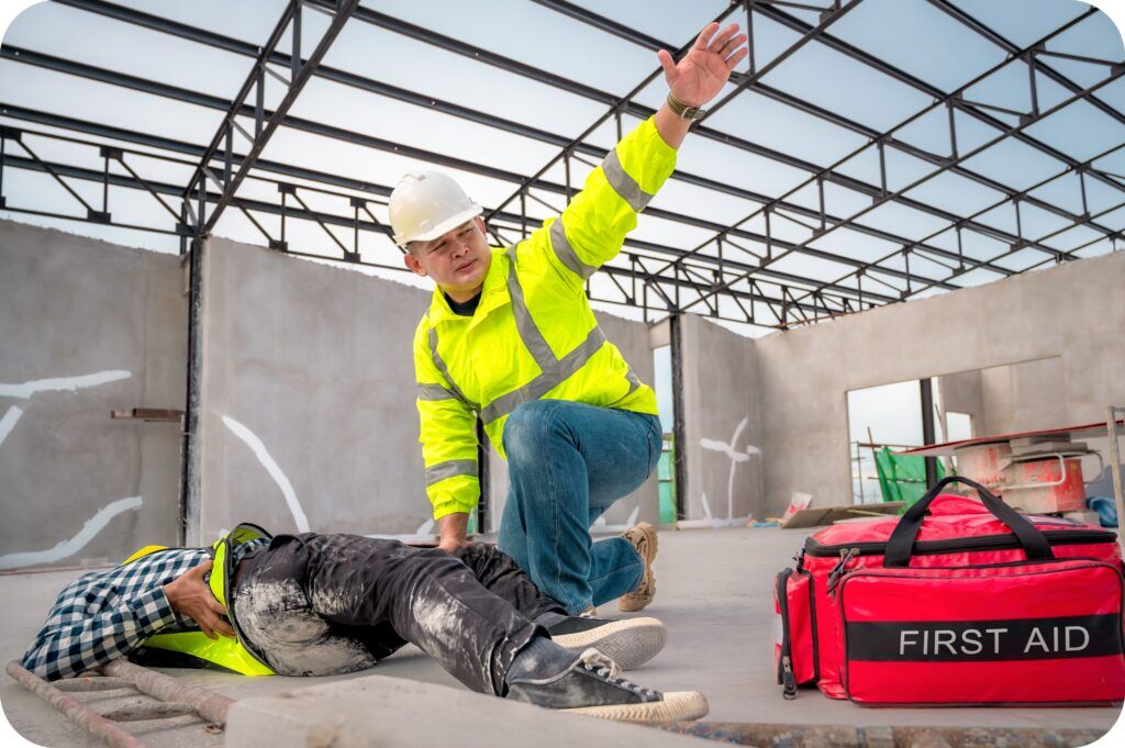 Worker kneeling to treat an injured coworker’s leg with a first aid kit nearby on a construction site.