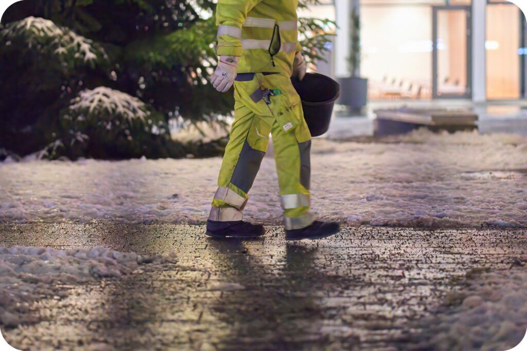Maintenance staff applying ice melt on a slick pathway at night to improve traction and prevent injuries.